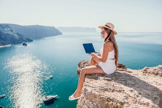 Successful business woman in yellow hat working on laptop by the sea. Pretty lady typing on computer at summer day outdoors. Freelance, travel and holidays concept.