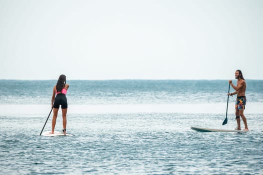 Sea woman and man on sup. Silhouette of happy young woman and man, surfing on SUP board, confident paddling through water surface. Idyllic sunset. Active lifestyle at sea or river