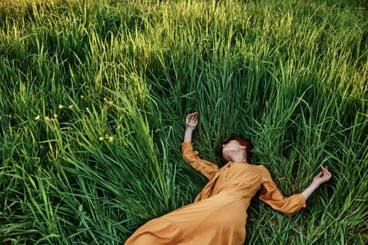 a sweet, calm woman in an orange dress lies in a green field with her arms outstretched and her eyes closed, enjoying the silence and peace. Horizontal photo taken from above. High quality photo