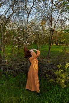 an elegant, sophisticated woman poses relaxed standing near a wicker fence at the dacha in a long orange dress tossing her long red hair. High quality photo