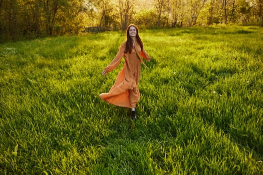 an attractive, slender, red-haired woman stands in a wide, green field during sunset in a long orange dress enjoying unity with nature and relaxation, raising her arms to the sides while facing the camera. High quality photo