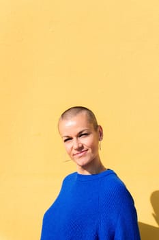 woman with a shaved head and a blue sweater smiling looking at camera with a yellow wall in the background, copy space for text