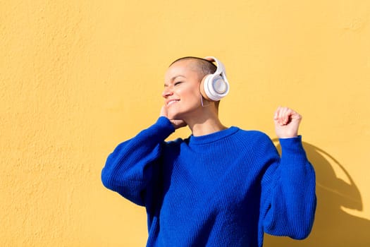 woman with a shaved head and a blue jumper dancing happy listening to music in her headphones with a yellow wall in the background, concept of rhythm and positive people, copy space for text