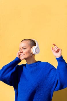 woman with a shaved head and a blue jumper dancing happy listening to music in her headphones with a yellow wall in the background, concept of rhythm and positive people, copy space for text