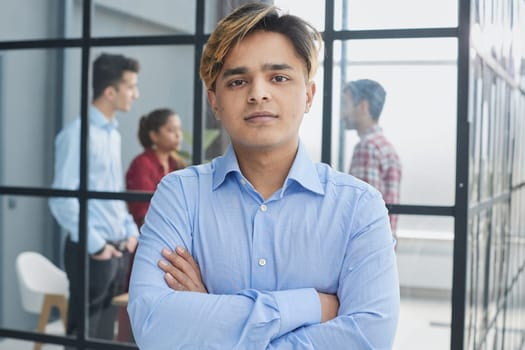 Portrait of a confident young businessman posing with his arms crossed in an office with his colleagues in the background.