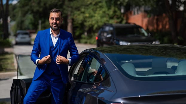 Caucasian bearded man in a blue suit gets out of a black electro car in the countryside in summer