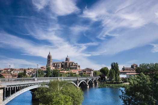 Perspective view of the bridge of Enrique Estevan over the Tormes river and the cathedral of Salamanca in the background with blue sky and light white clouds.