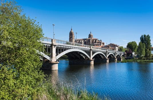 Perspective view of the bridge of Enrique Estevan over the Tormes river and the cathedral of Salamanca in the background with clear blue sky.