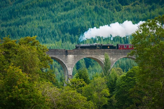 Old steam train crossing the Glen Viaduct surrounded by vegetation in Scotland. Jacobite steam train.