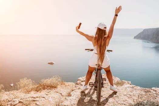 A woman cyclist on a mountain bike looking at the landscape of mountains and sea. Rear view of cyclist woman standing in front to the sea with outstretched arms. Freedom and healthy lifestyle concept