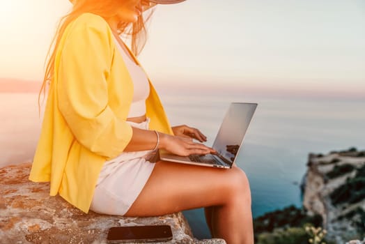 Successful business woman in yellow hat working on laptop by the sea. Pretty lady typing on computer at summer day outdoors. Freelance, travel and holidays concept.