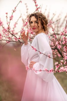 Woman peach blossom. Happy curly woman in white dress walking in the garden of blossoming peach trees in spring.