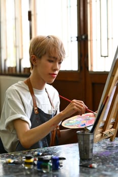 Portrait of teenage man painting picture with watercolor on canvas in art workshop. Art, creative hobby and leisure activity concept.