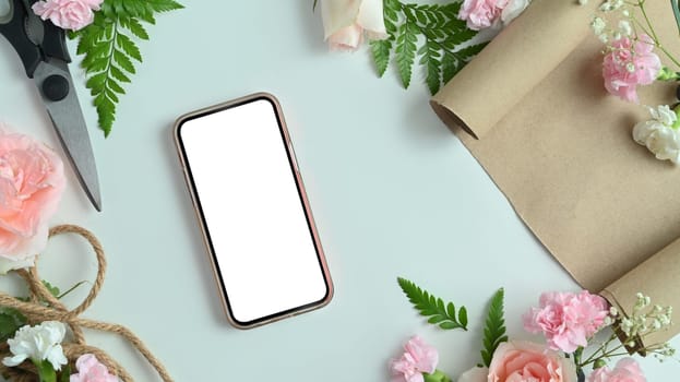 Smartphone with blank screen, pink flowers, shears, wrapping paper and leaves arrangement on white background.