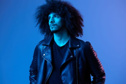 Portrait of fashion man with curly hair on blue background multinational, colored light, black leather jacket trend, modern concept. High quality photo