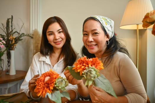 Image of adult daughter and middle age mother holding bouquet of orange flowers and smiling to camera. Happy mother's day.
