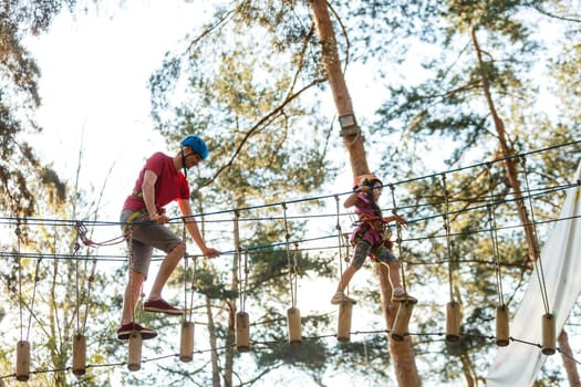 Young father teaching her daughter to climb on the rock climbing wall. Little girl preschooler wearing safety harness having fun time in adventure rope park. Happy family concept.