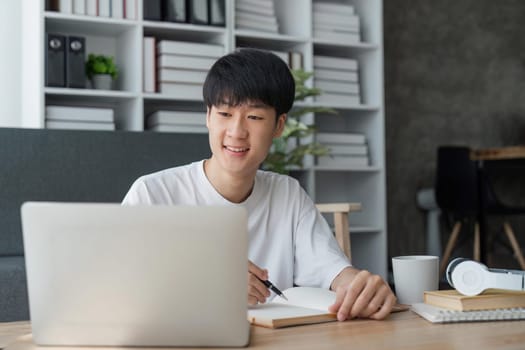 young man studying at home using his laptop, receiving lectures online and taking notes, smiling.