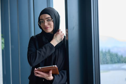 A hijab businesswoman stands by the window in a modern office holding a tablet in her hand showcasing her professionalism, technological prowess and entrepreneurial spiri.