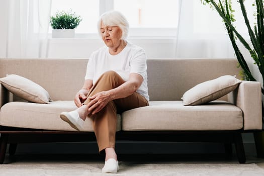 Elderly woman severe pain in her leg sitting on the couch, health problems in old age, poor quality of life. Grandmother with gray hair holds on to her sore knee, problems with joints and ligaments. . High quality photo
