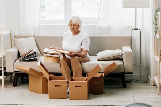 elderly woman sits on a sofa at home with boxes. collecting things with memories albums with photos and photo frames moving to a new place cleaning things and a happy smile. Lifestyle retirement. High quality photo