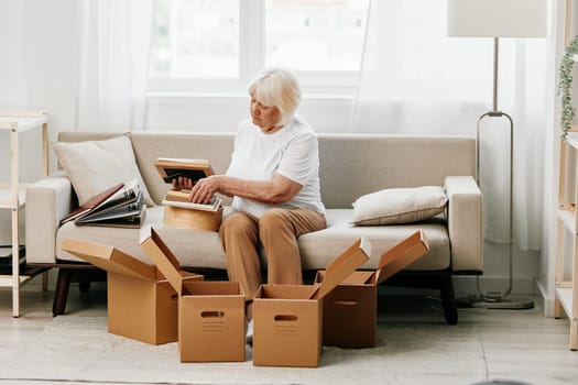 elderly woman sits on a sofa at home with boxes. collecting things with memories albums with photos and photo frames moving to a new place cleaning things and a happy smile. Lifestyle retirement. High quality photo