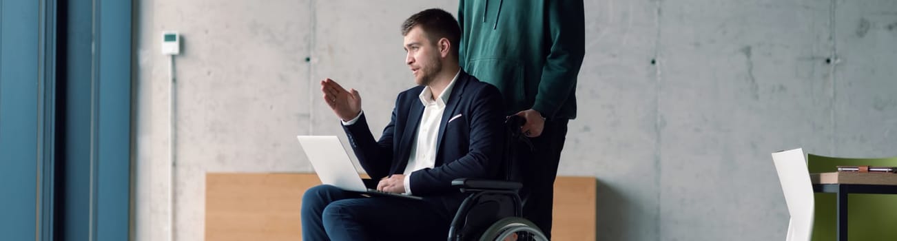 Wide crop photo of businessman in a wheelchair in a fashionable office using a laptop while behind him is his business colleague who gives him support.