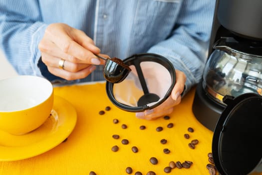 A young woman prepares herself coffee in a bright yellow mug. Grains of coffee on the table, against the background of a drip coffee maker. Enjoy fresh aromatic coffee