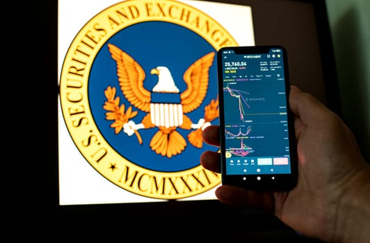 Kyiv, Ukraine - 06 June 2023: Man holds a smartphone with falling Bitcoin chart on Binance against the background the SEC logo. S.E.C. Accuses Binance of Mishandling Funds and Lying to Regulators