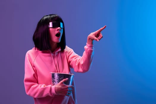 Excited awesome brunet woman in pink hoodie trendy specular sunglasses with popcorn point finger aside posing isolated in blue violet color light background. Neon party Cinema concept. Copy space
