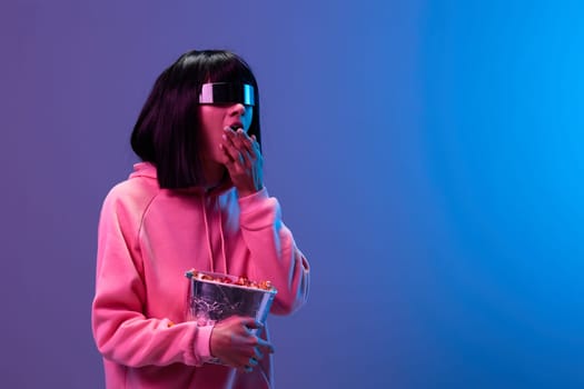 Shocked overjoyed awesome brunet woman in pink hoodie trendy specular sunglasses eat popcorn look aside posing isolated in blue violet color light background. Neon party Cinema concept. Copy space