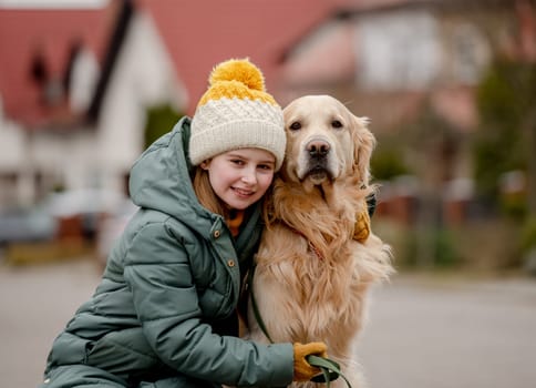Preteen child girl hugging golden retriever dog at autumn city street wearing hat and warm jacket. Pretty kid with purebred pet doggy labrador outdoors