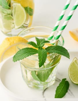 Lemonade in a transparent glass with lemon, lime, rosemary sprigs and mint leaves on a white background