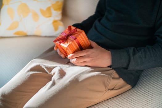 A funny erotic gift for a partner for a holiday, anniversary, Valentine's day, a young man sits on the couch and holds a small gift wrapped in a box, tied with a ribbon in an intimate place.