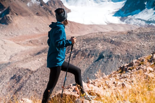 A young girl leads an active lifestyle, climbs a trail in the mountains with trekking poles, snow-capped mountain peaks and a glacier in the background.