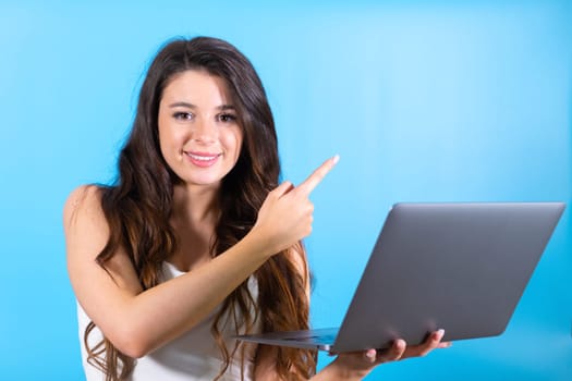Long-haired woman with delighted and amused expression looks at ad news on laptop screen. Lady points to advertised modern boutique at blue wall
