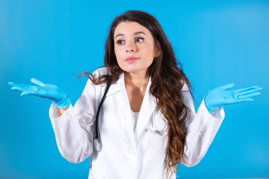 Brown-haired doctor in gloves and with stethoscope looks around for patient at blue office wall. Female nurse looks with confused and concerned expression