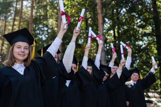 A group of graduates in robes raised their hands with diplomas outdoors. Elderly student