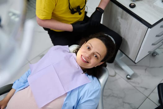 Portrait of multi-ethnic young pregnant woman expecting baby, sitting in dental chair, smiling with a beautiful smile, looking at camera during a dentist check up in dentistry clinic. View from above