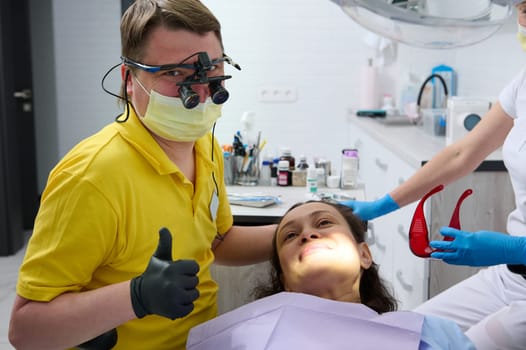 Happy professional dentist, hygienist orthodontist in protective medical mask and goggles, looking at camera while performing oral examination of female patient in dentistry clinic. Dental practice