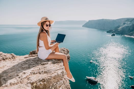 Successful business woman in yellow hat working on laptop by the sea. Pretty lady typing on computer at summer day outdoors. Freelance, travel and holidays concept.