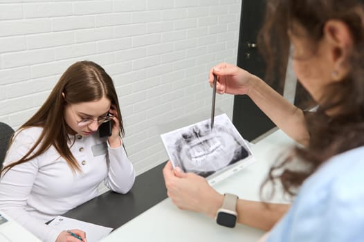 Female receptionist talks on mobile phone. Blurred foreground of a dentist holding dental panoramic x-ray and explaining to his patient on all the dental treatment that needs to do in dentistry clinic