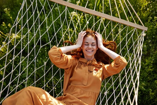 a beautiful, elegant woman lies in a long orange dress on a mesh hammock resting in nature, illuminated by the warm sunset light, smiling happily holding her hands near her face. High quality photo