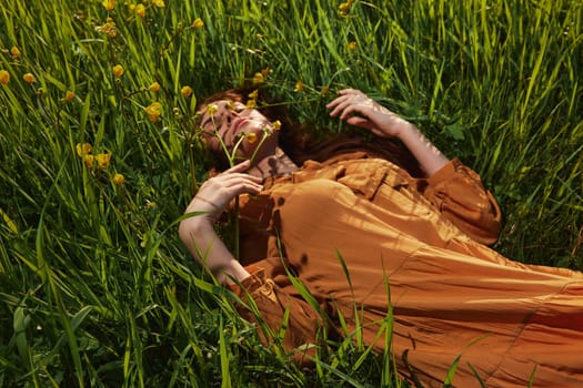 a calm woman with long red hair lies in a green field with yellow flowers, in an orange dress with her eyes closed, with a pleasant smile on her face, enjoying peace and recuperating, illuminated by the setting sun. High quality photo