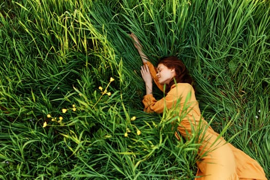 a relaxed woman enjoys summer lying in the tall green grass in a long orange dress stretched out stretching her arm forward. High quality photo