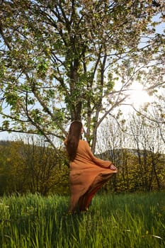 a slender, attractive woman with long red hair is standing in the countryside near a flowering tree in a long orange dress and happily spinning holding the dress. Vertical photography. High quality photo