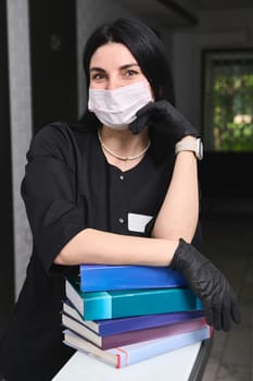 Authentic portrait of a beautiful experienced female doctor dentist wearing stylish black uniform, medical mask and gloves, standing with a pile of medical literature, looking confidently at camera