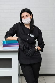 Confident female doctor intern in black medical uniform and mask, looks at camera, standing with a stack of medical literature over white brick wall background in a medical clinic. Medicine. Education