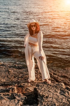 woman sea sunset. woman in a white pantsuit and hat is standing on the beach enjoying the sea. Happy summer holidays.