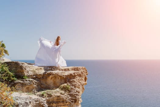 Woman in a white dress on the sea. Side view Young beautiful sensual woman in white long dress posing on a rock high above the sea at sunset. Girl in nature against the blue sky.